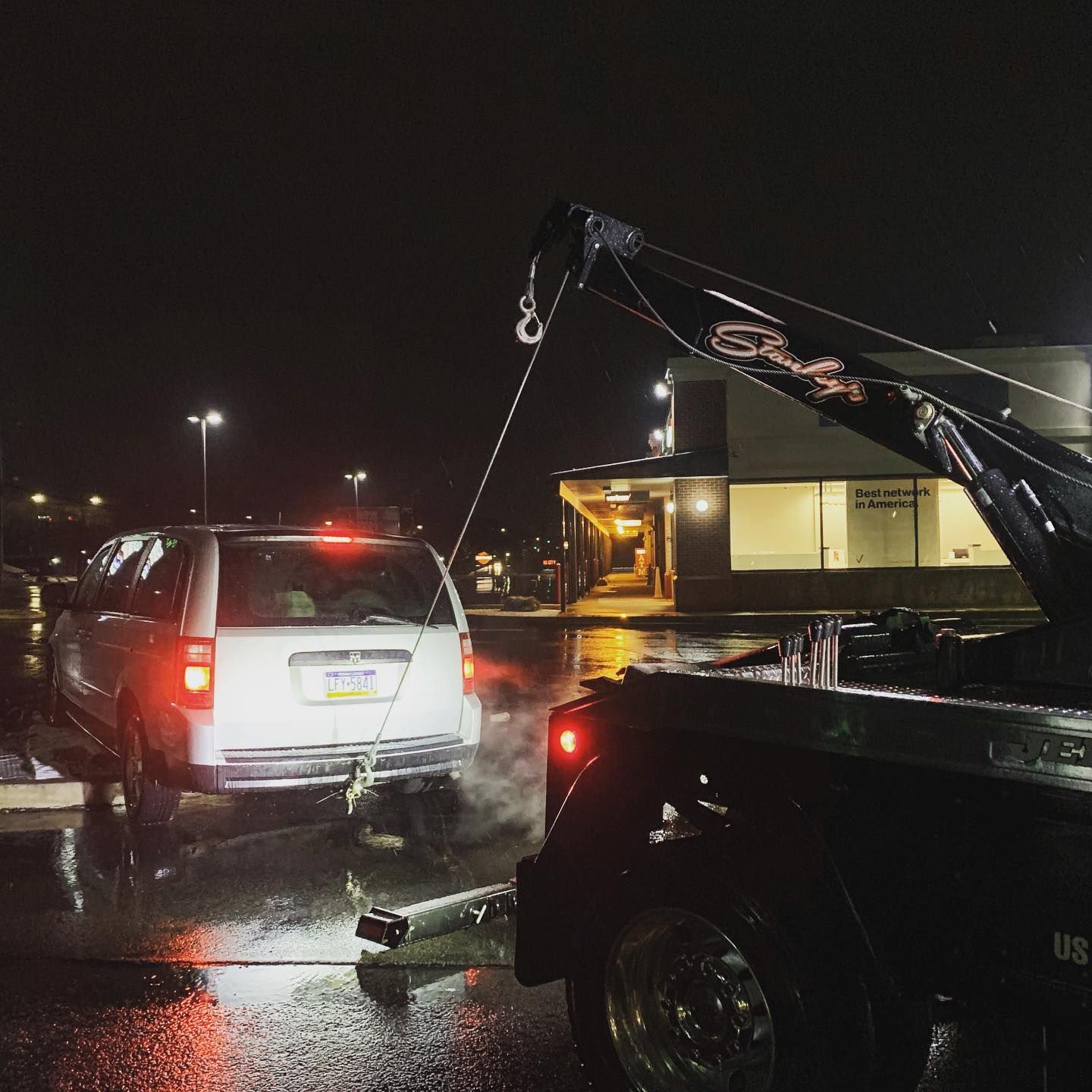 A tow truck pulling a white minivan in a wet parking lot at night.