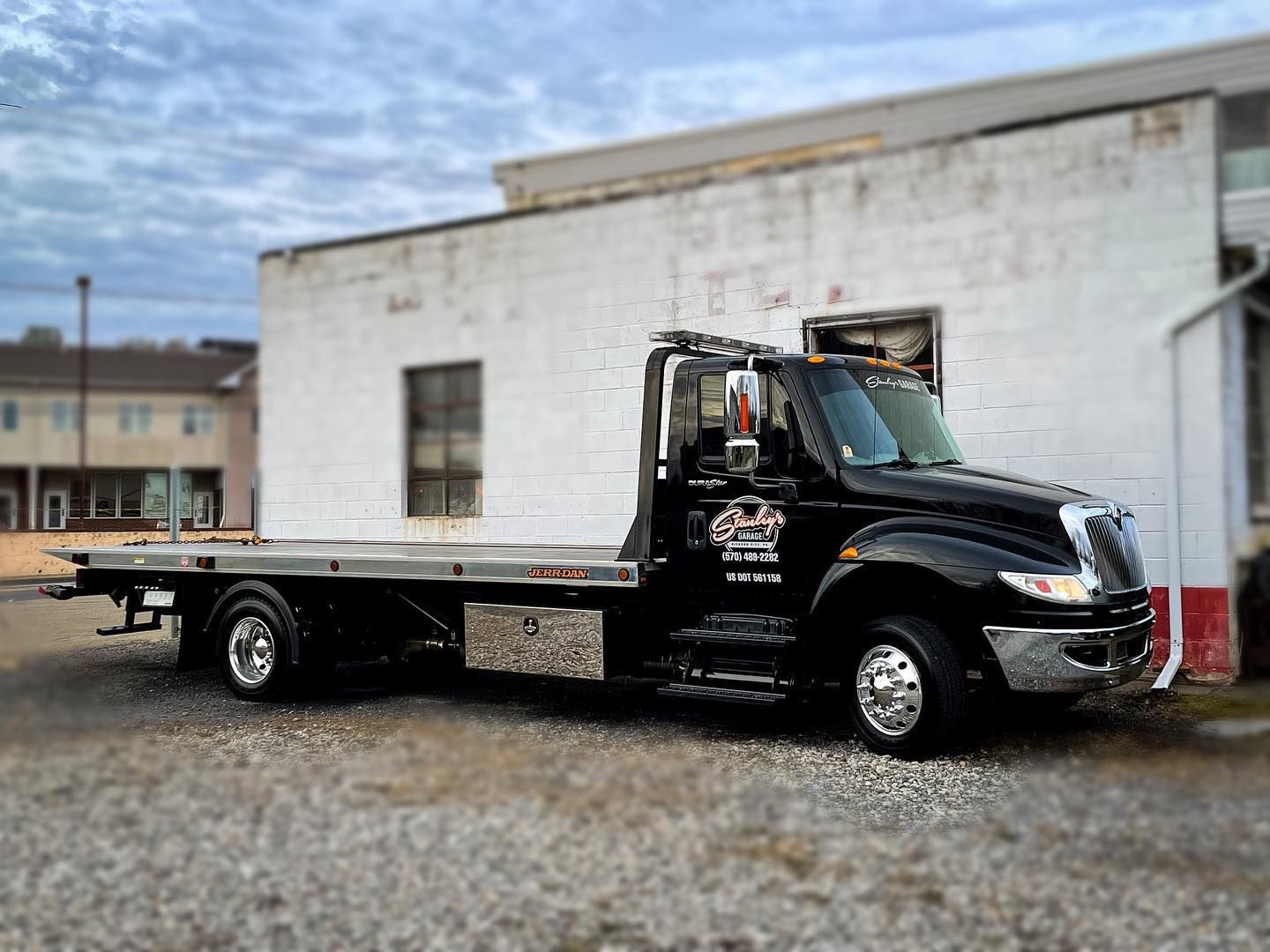 Black tow truck parked in front of a white building with cloudy sky.