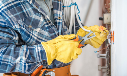 A man in a plaid shirt and yellow gloves is working on an electrical outlet.