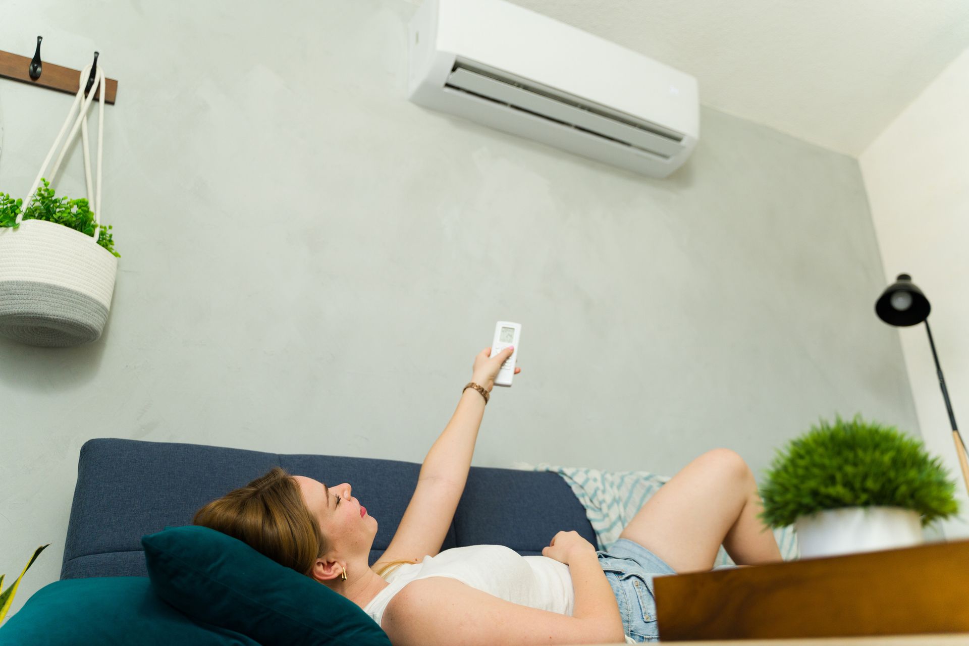 Young woman on sofa using remote, enjoying cool air from AC on hot summer day.