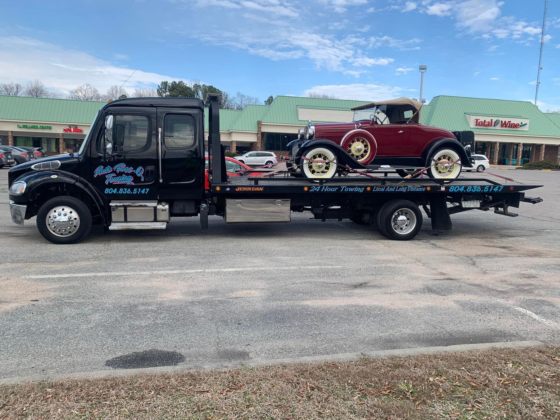 Black tow truck with vintage maroon car, parked in a lot. Cloudy sky, shopping center in the background.