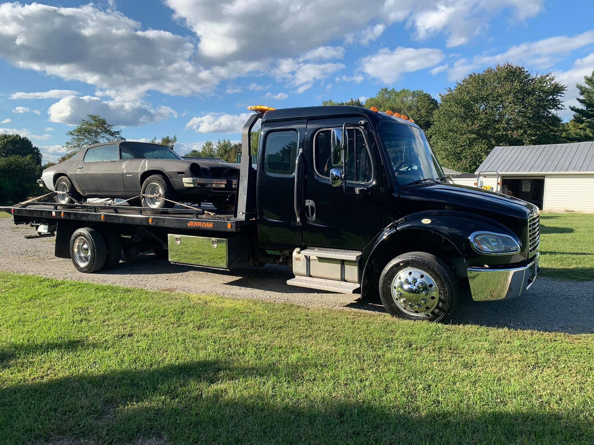 Black tow truck carrying a silver car on a grassy area under a cloudy sky.