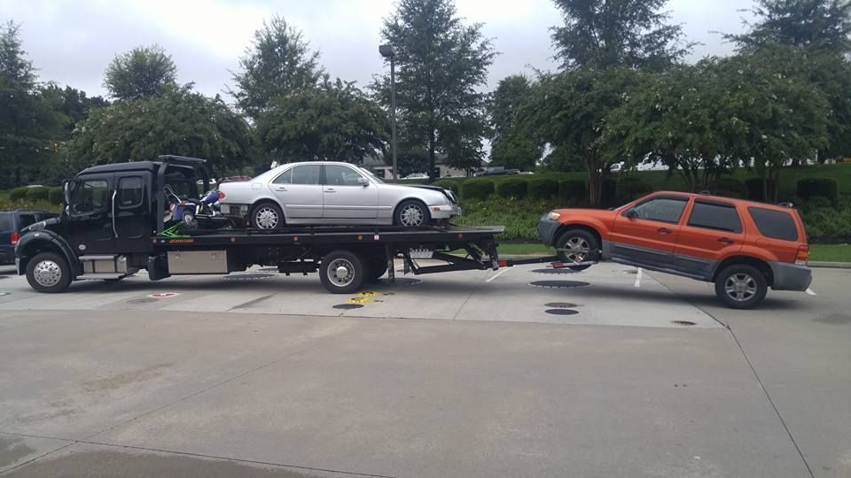 Tow truck carrying a silver sedan and an orange SUV on a flatbed in a parking lot.
