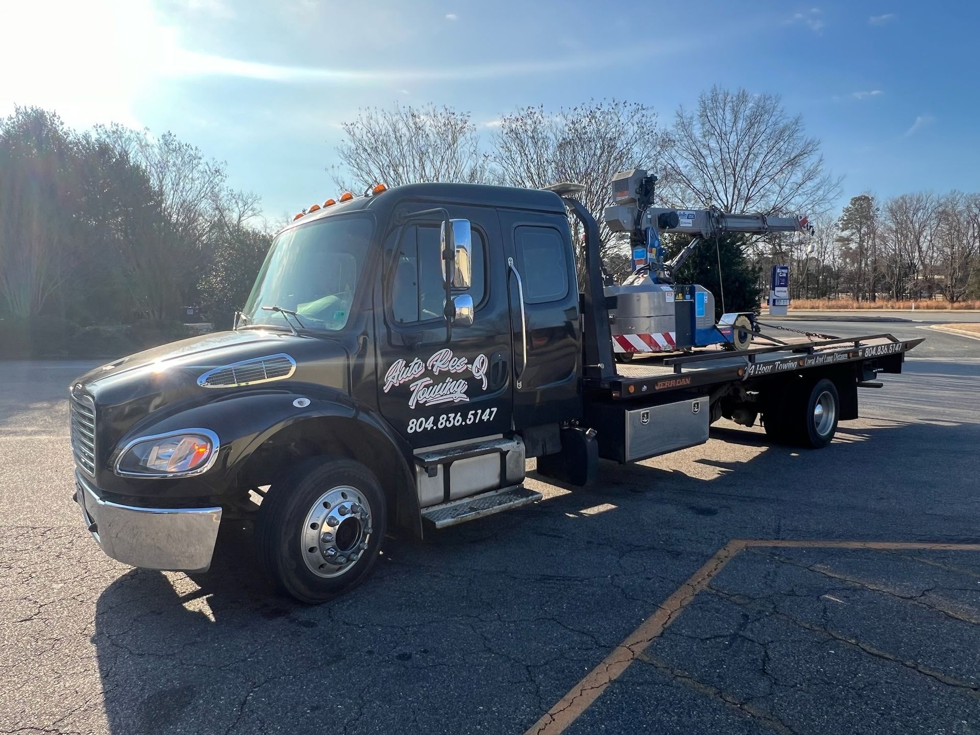 Black tow truck parked on asphalt; bright sunlight, trees in background.