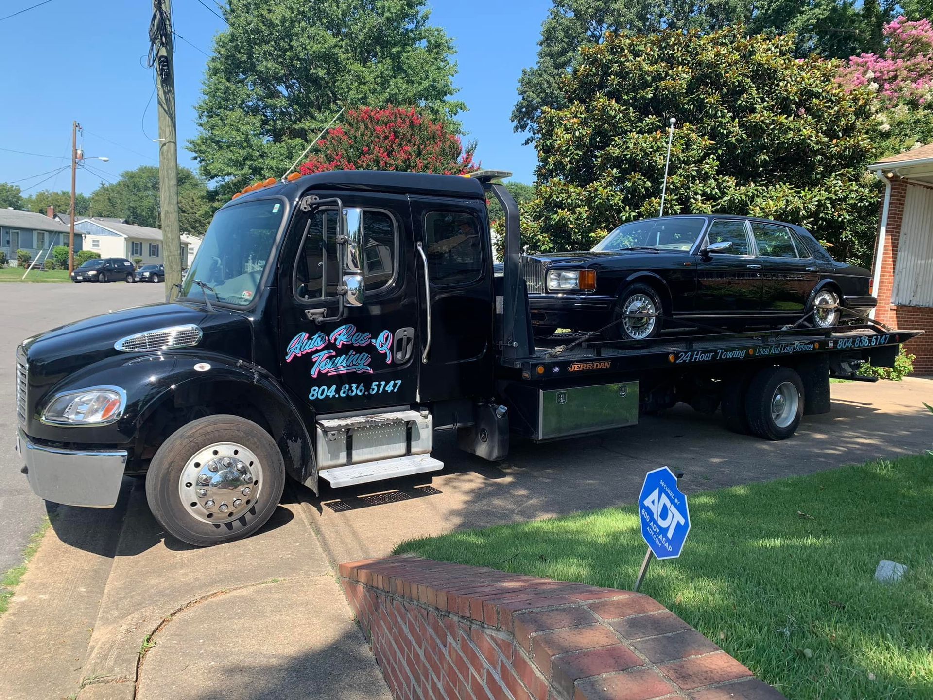 Black tow truck hauling a black sedan on a sunny residential street.