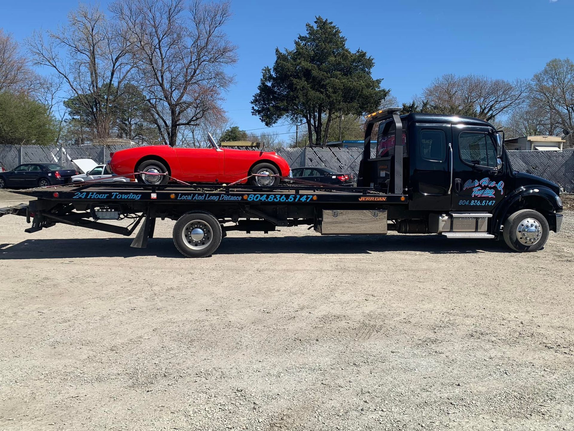 A red convertible car loaded on a black tow truck on a gravel lot under a blue sky.