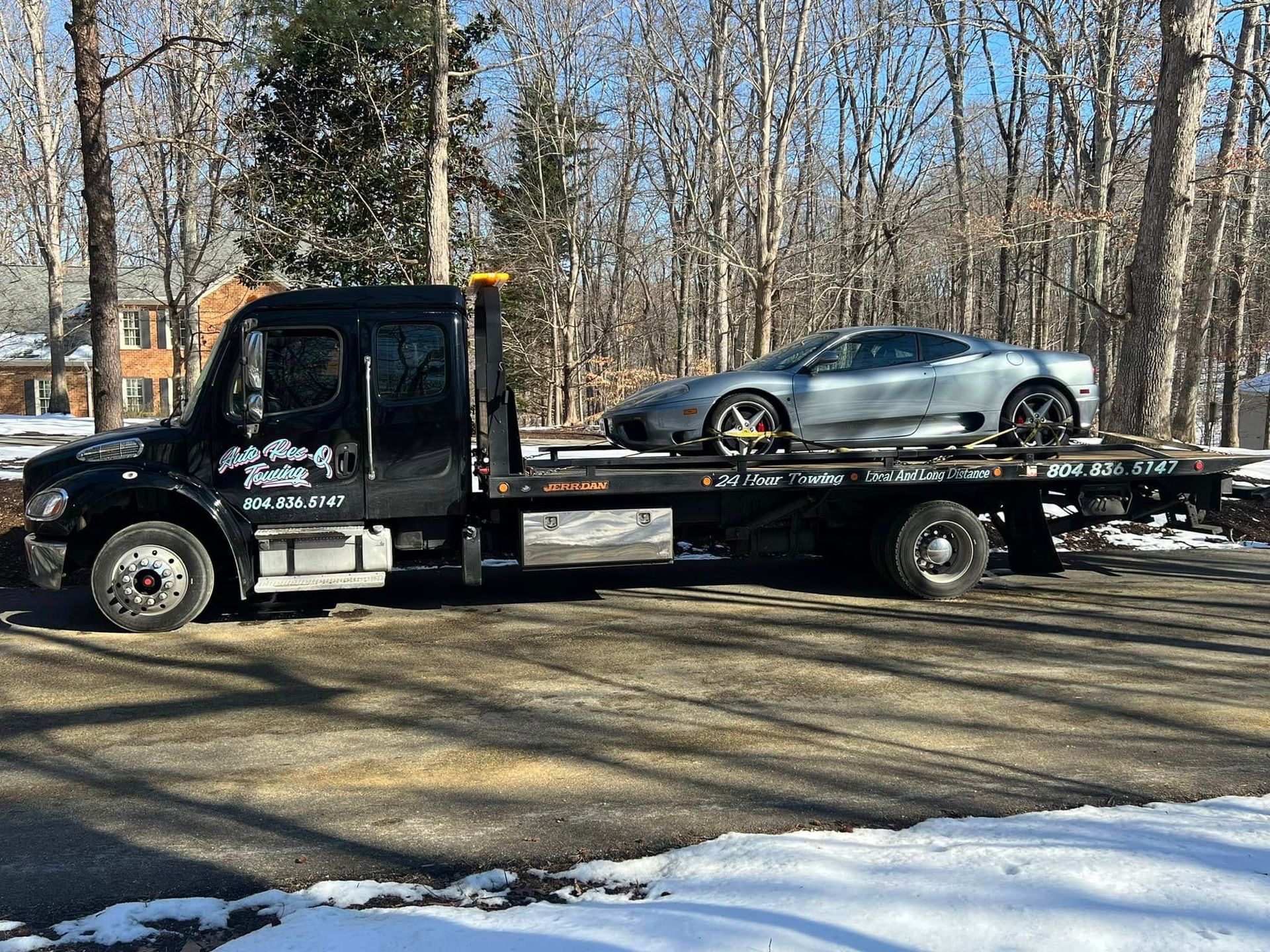 Black tow truck carrying a silver sports car on a snowy day.