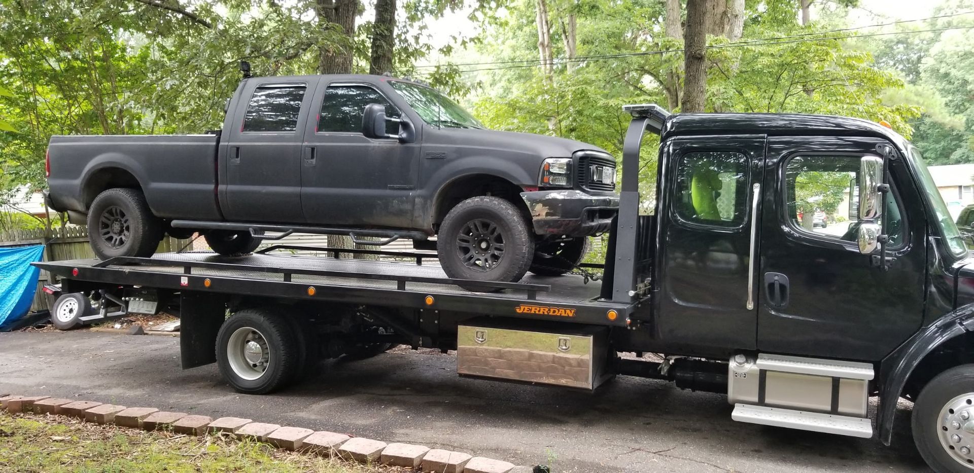 Black pickup truck on a black flatbed tow truck on a paved driveway; trees in the background.