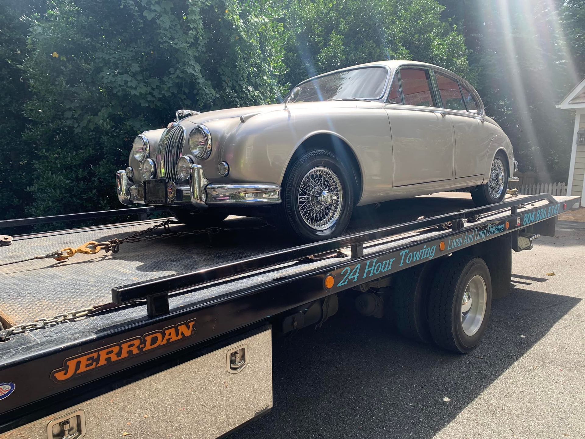 A classic silver Jaguar sedan is secured on a flatbed tow truck outdoors on a sunny day.