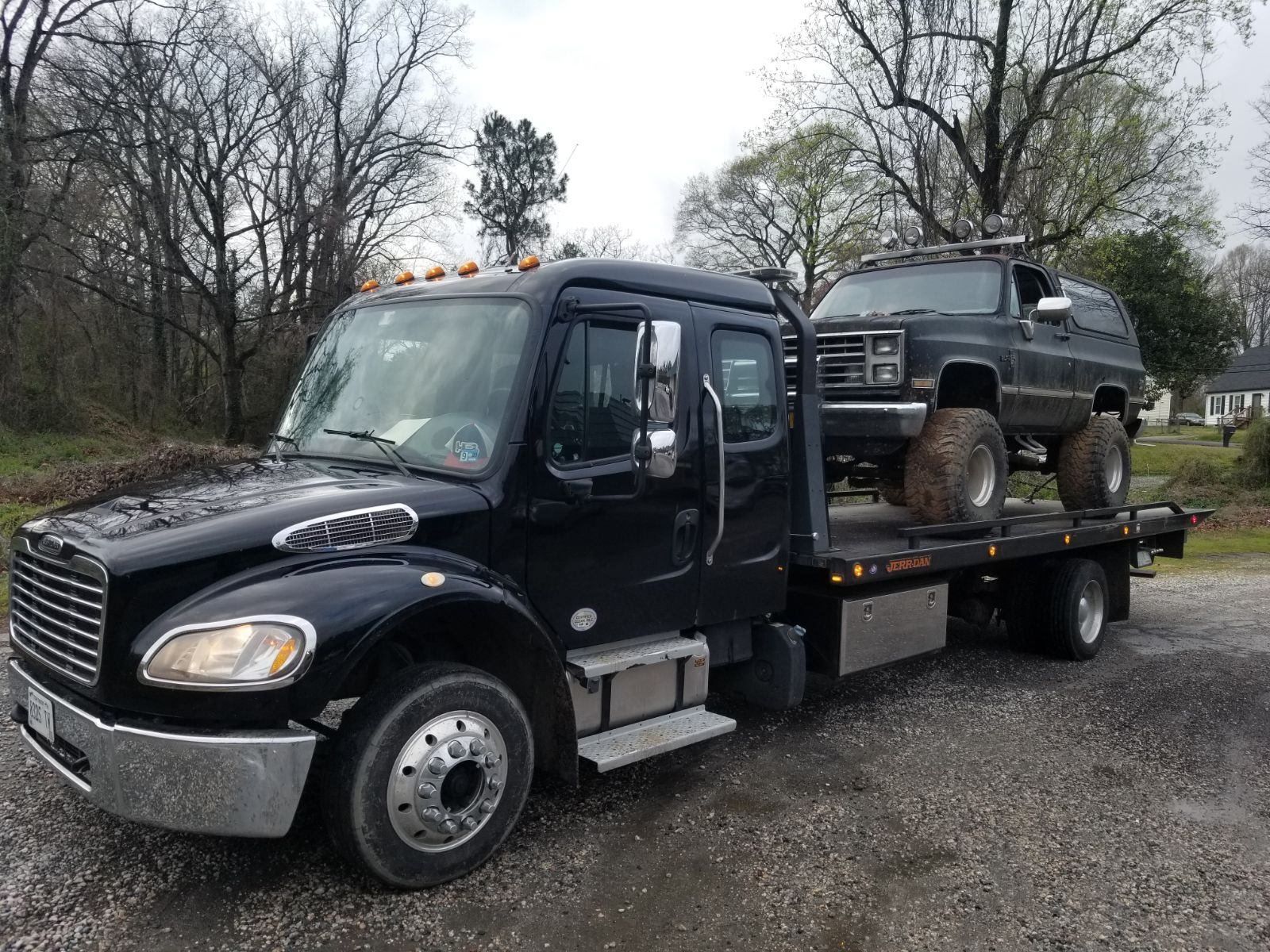 Black tow truck hauling a gray lifted SUV outdoors on a cloudy day.