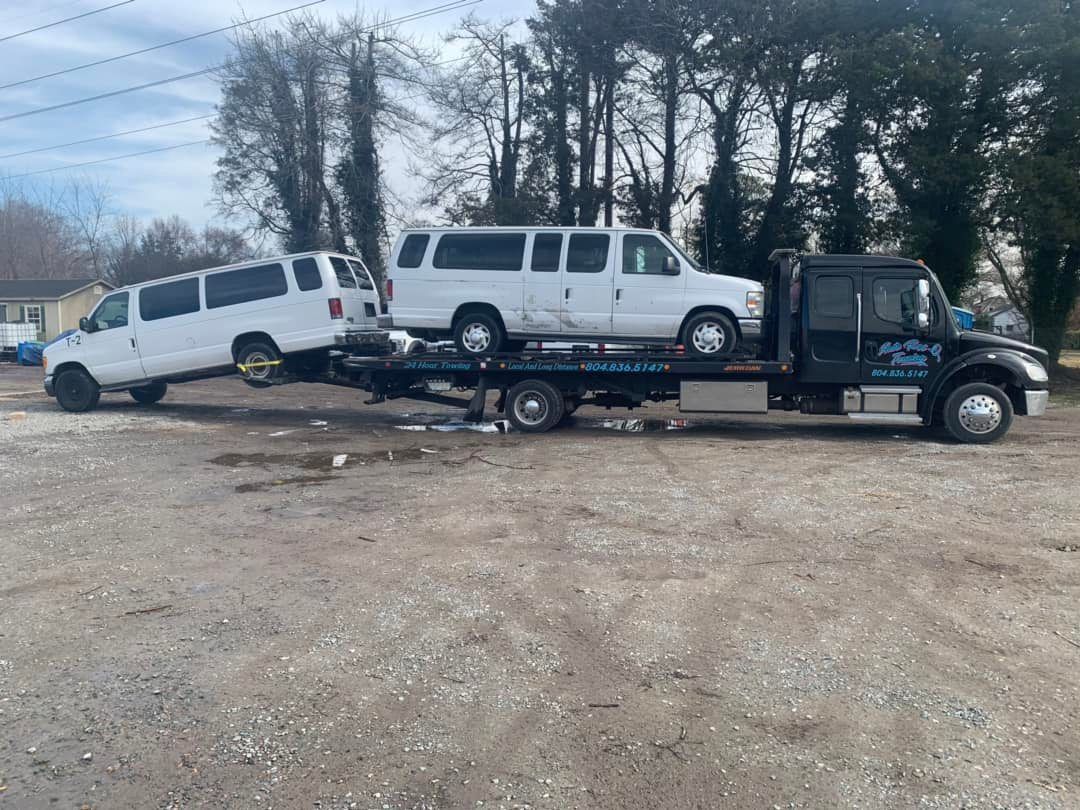 Two white vans being towed by a black tow truck on a gravel lot. One van is on the tow truck.