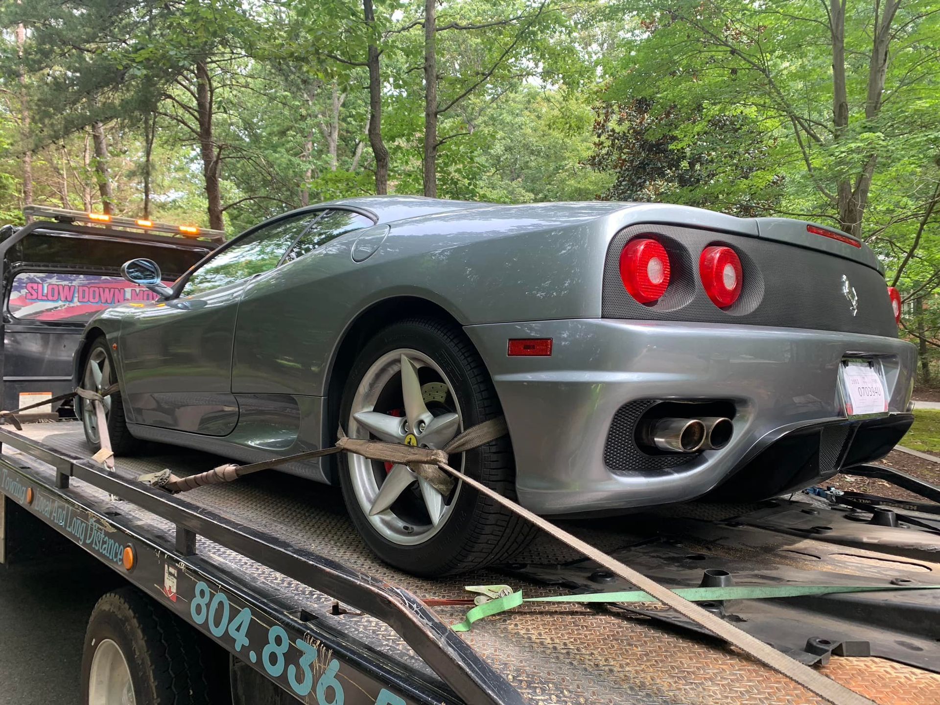 Silver Ferrari sports car on a tow truck, being transported on a road surrounded by trees.