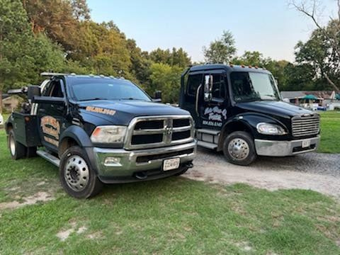 Two black tow trucks parked on grass, one Dodge, one Freightliner, in a residential area.