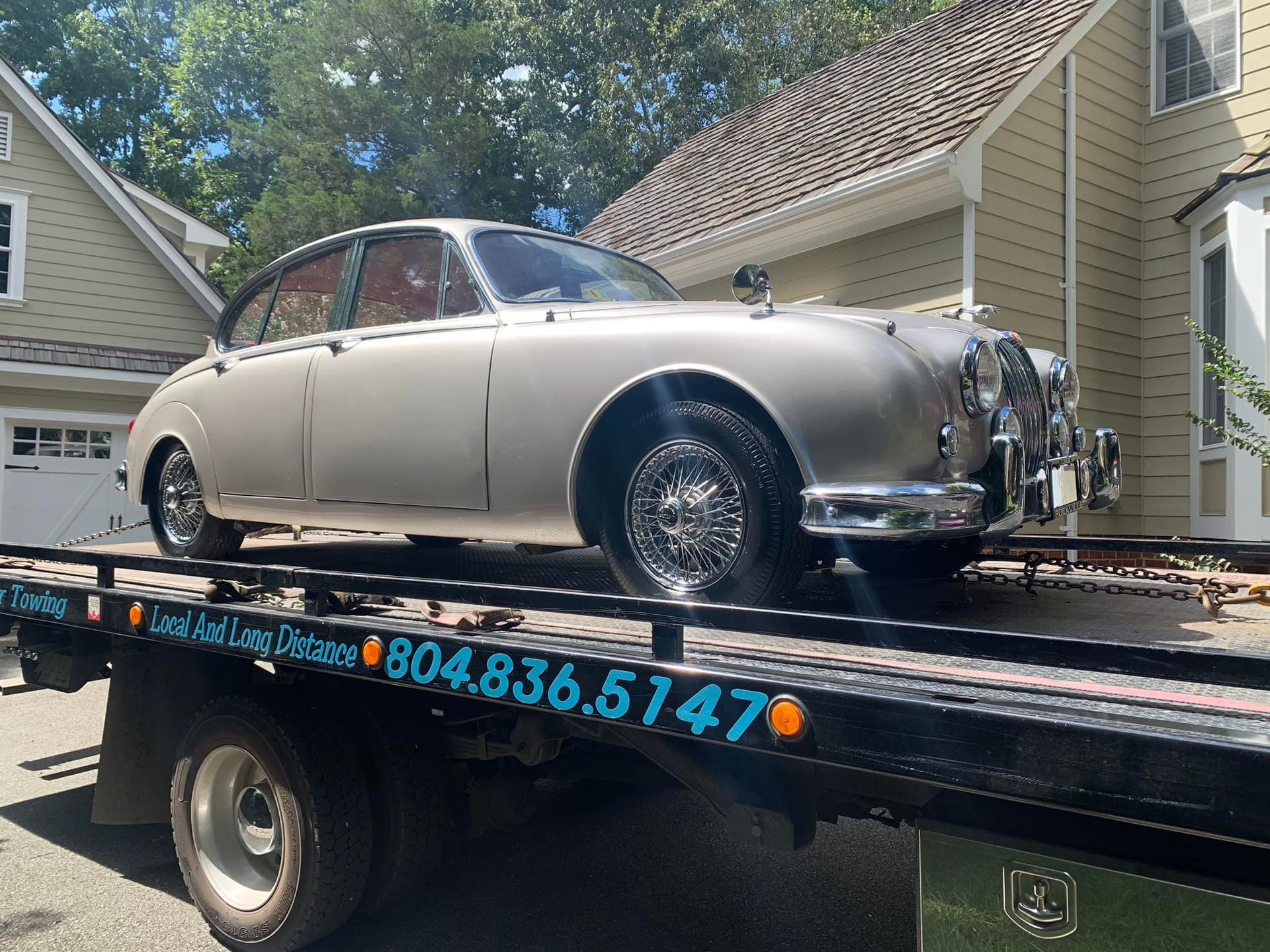 Silver vintage Jaguar sedan on a tow truck, parked in front of a house.