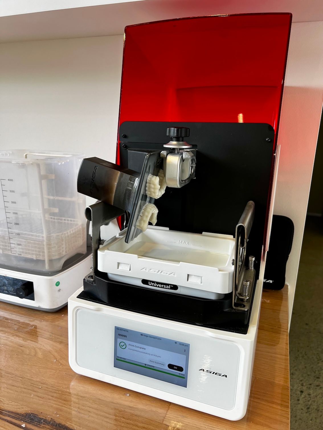 A white 3D resin printer with a red cover, displaying a print in progress. Next to it is a cleaning device— AB Esthetic Dental Laboratory in Robina, QLD