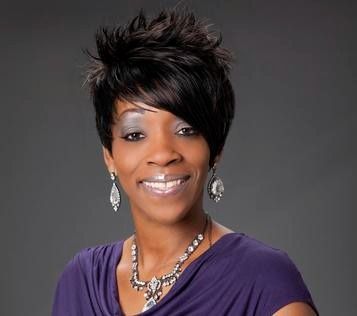 Woman with spiky black hair, wearing a purple shirt, silver necklace, and earrings, smiling against a gray background.