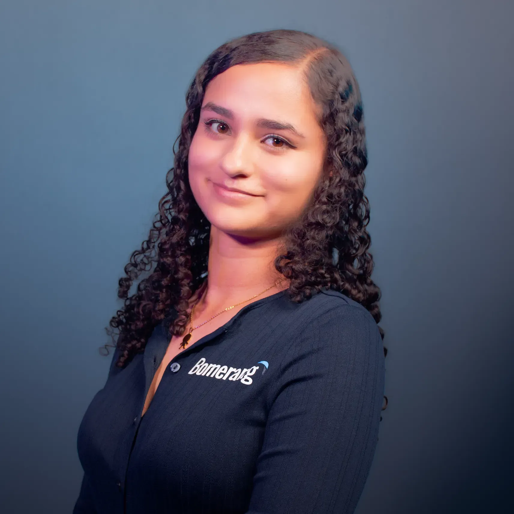 Una mujer con cabello rizado sonríe, vistiendo una camisa azul oscuro con logo, delante de un fondo azul.