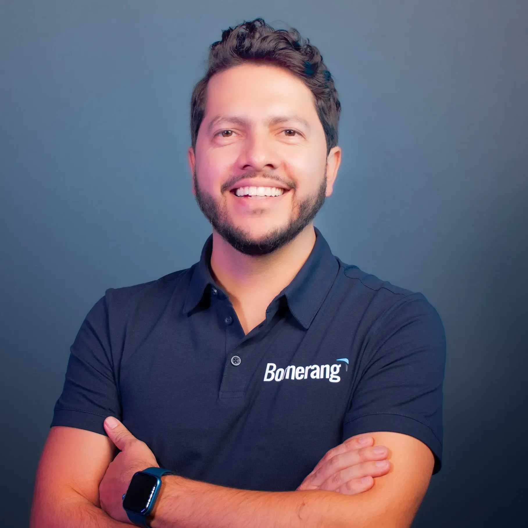 Hombre con los brazos cruzados sonriendo; vistiendo una camiseta polo negra con el logo 