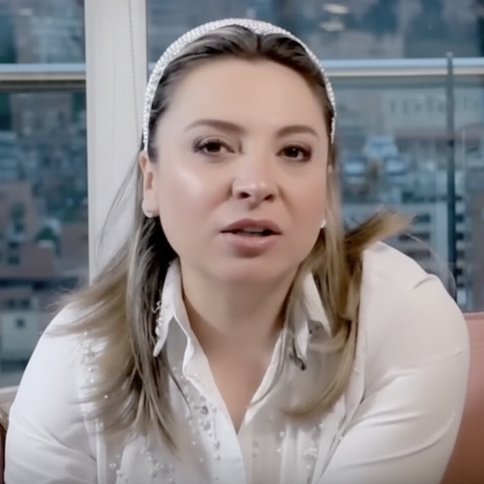 Mujer rubia con diadema y camisa blancas, hablando. Luz natural y vista de la ciudad al fondo.