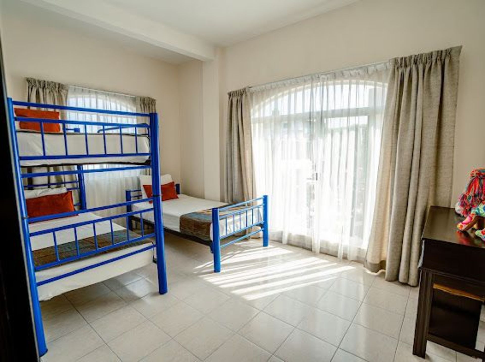 Habitación luminosa con una litera azul, una cama individual y una gran ventana con cortinas transparentes.