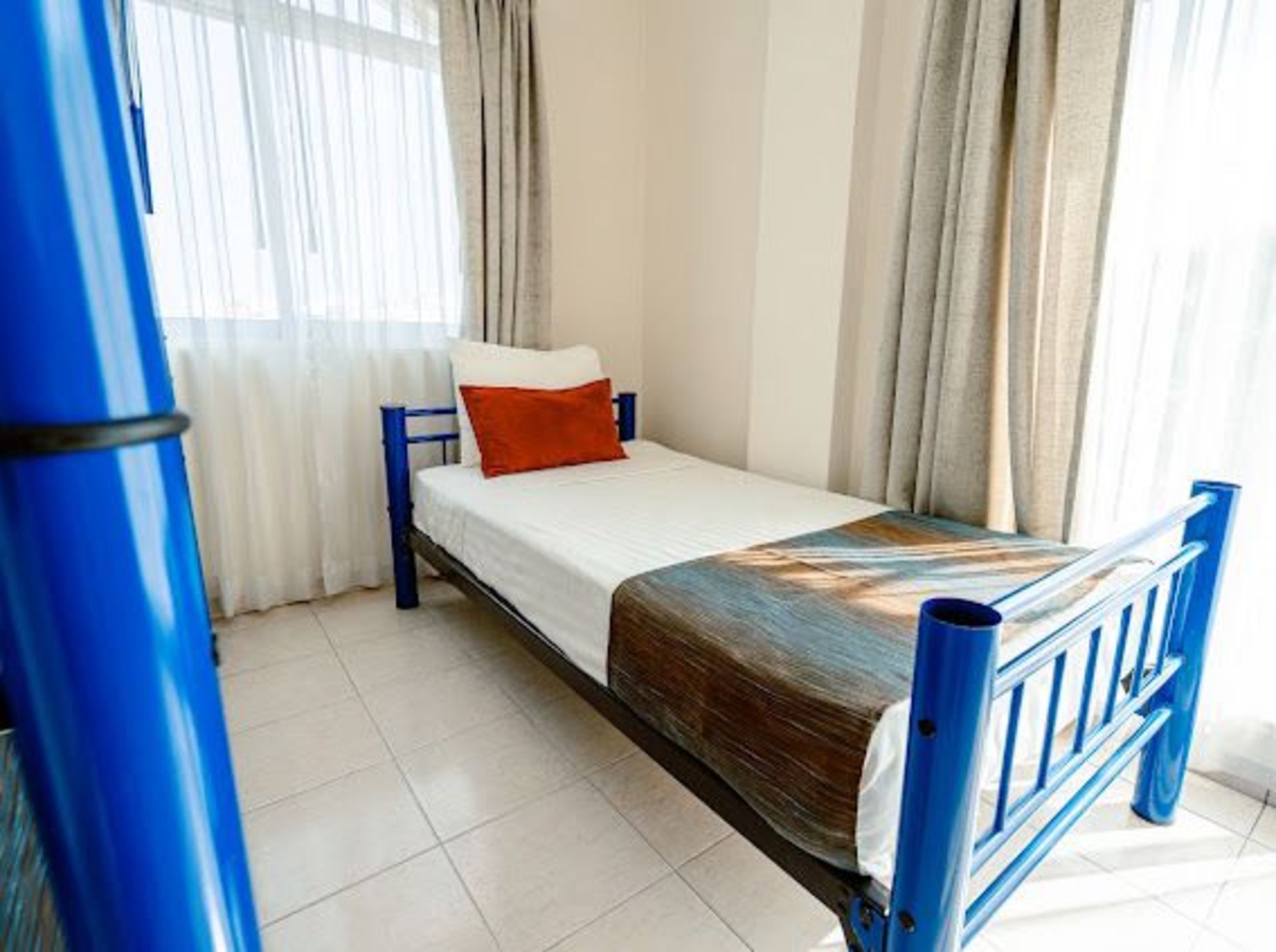 Una cama individual con marco azul, sábanas blancas y una almohada color óxido cerca de una ventana con cortinas.