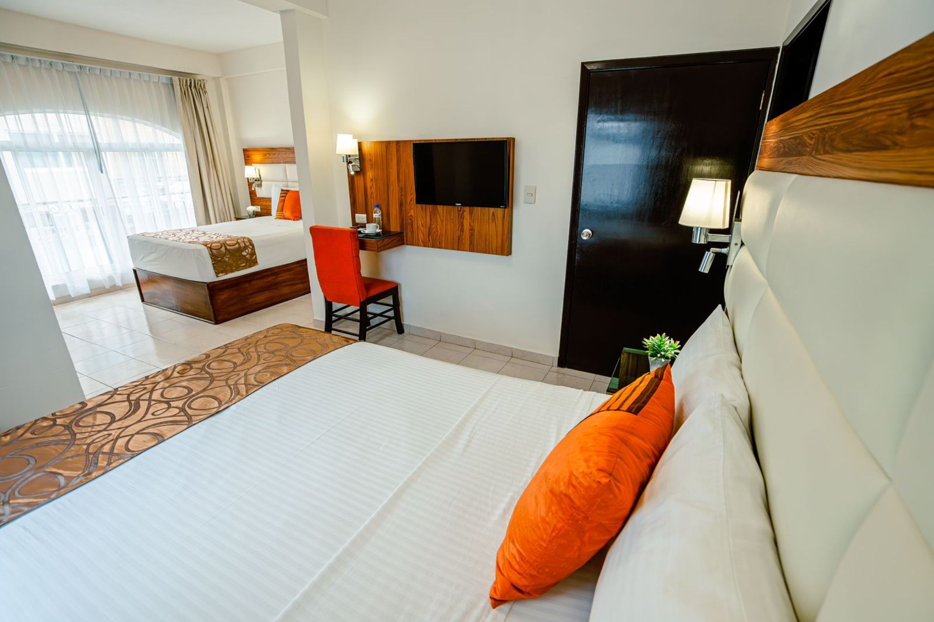 Habitación de hotel con cama king, escritorio y otra cama al fondo. Decoración moderna y luminosa con detalles en naranja.