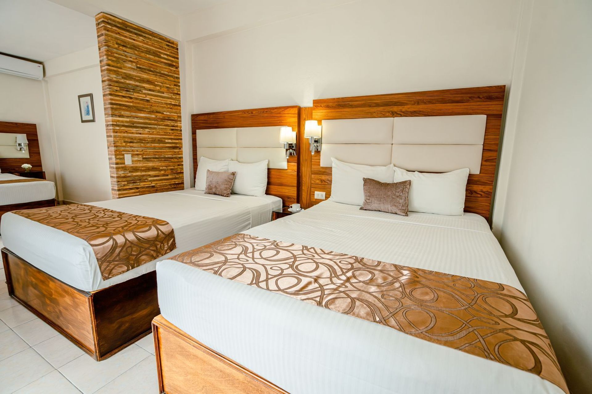 Habitación de hotel con dos camas queen, paredes de colores claros y cabeceros de madera, y un panel decorativo de madera.