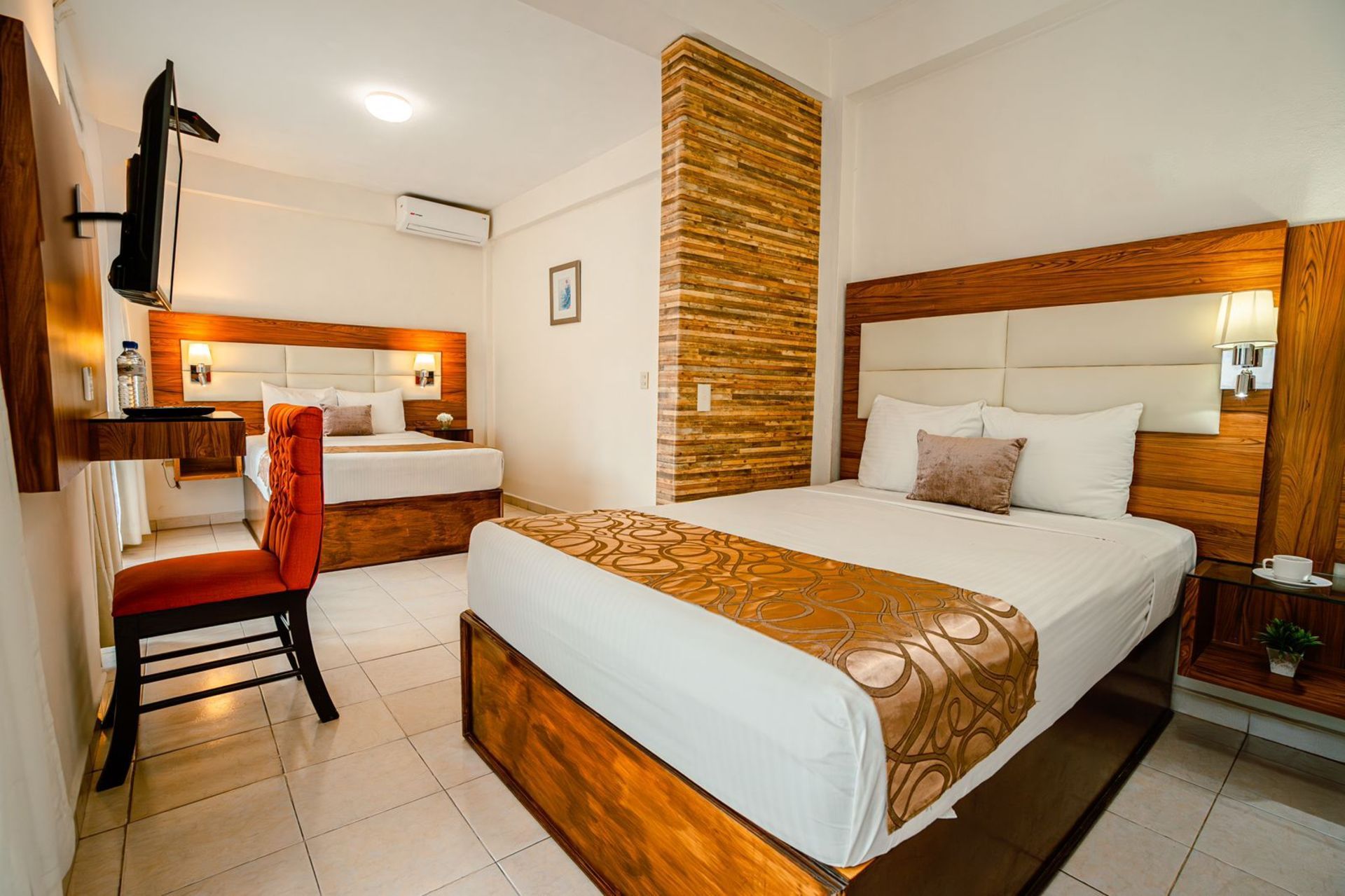 Habitación de hotel con dos camas, cabeceros de madera, una pequeña mesa con una silla y un televisor en la pared.