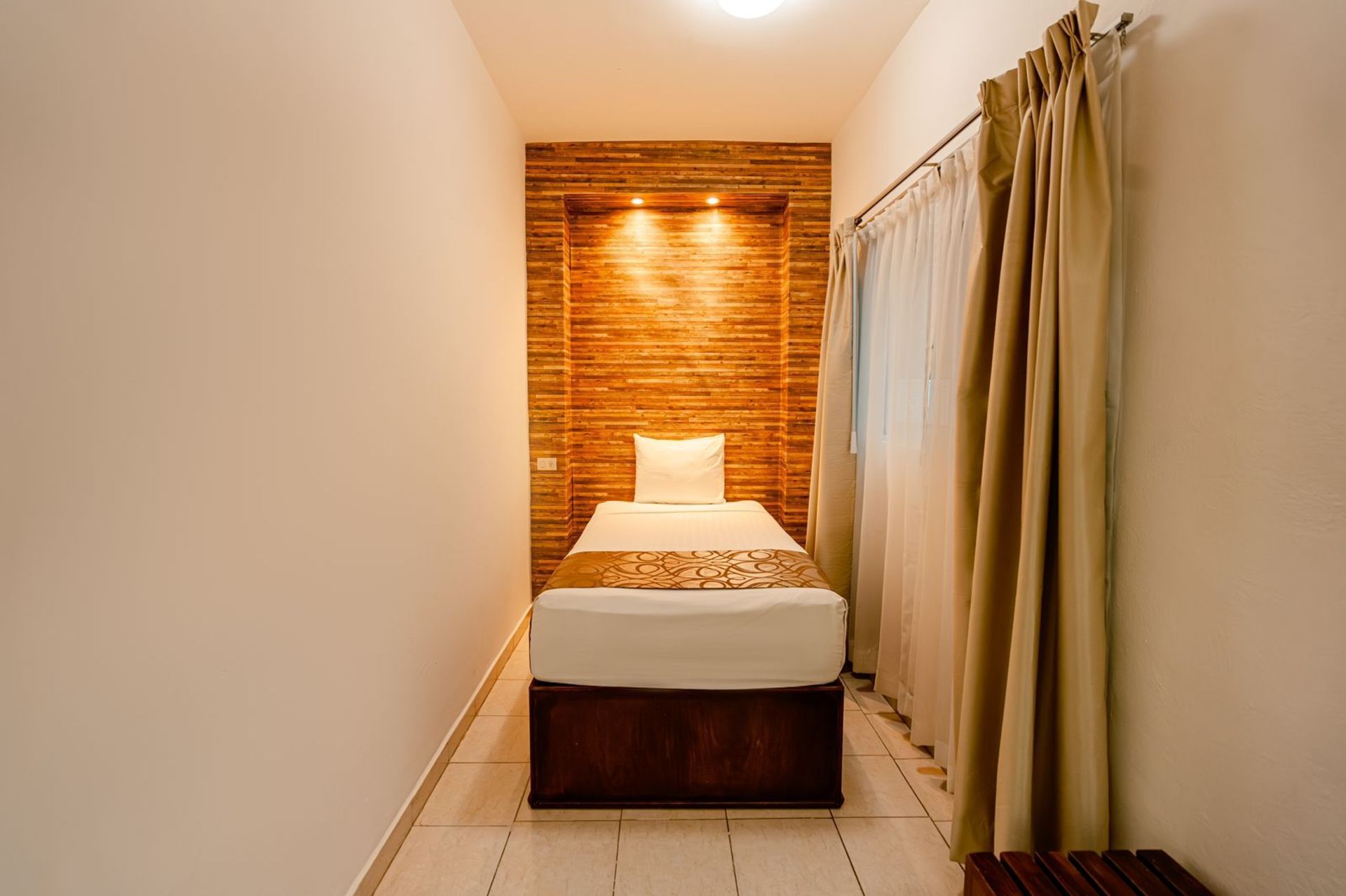 Habitación de hotel estrecha con una cama individual, pared con detalles de madera y cortinas.
