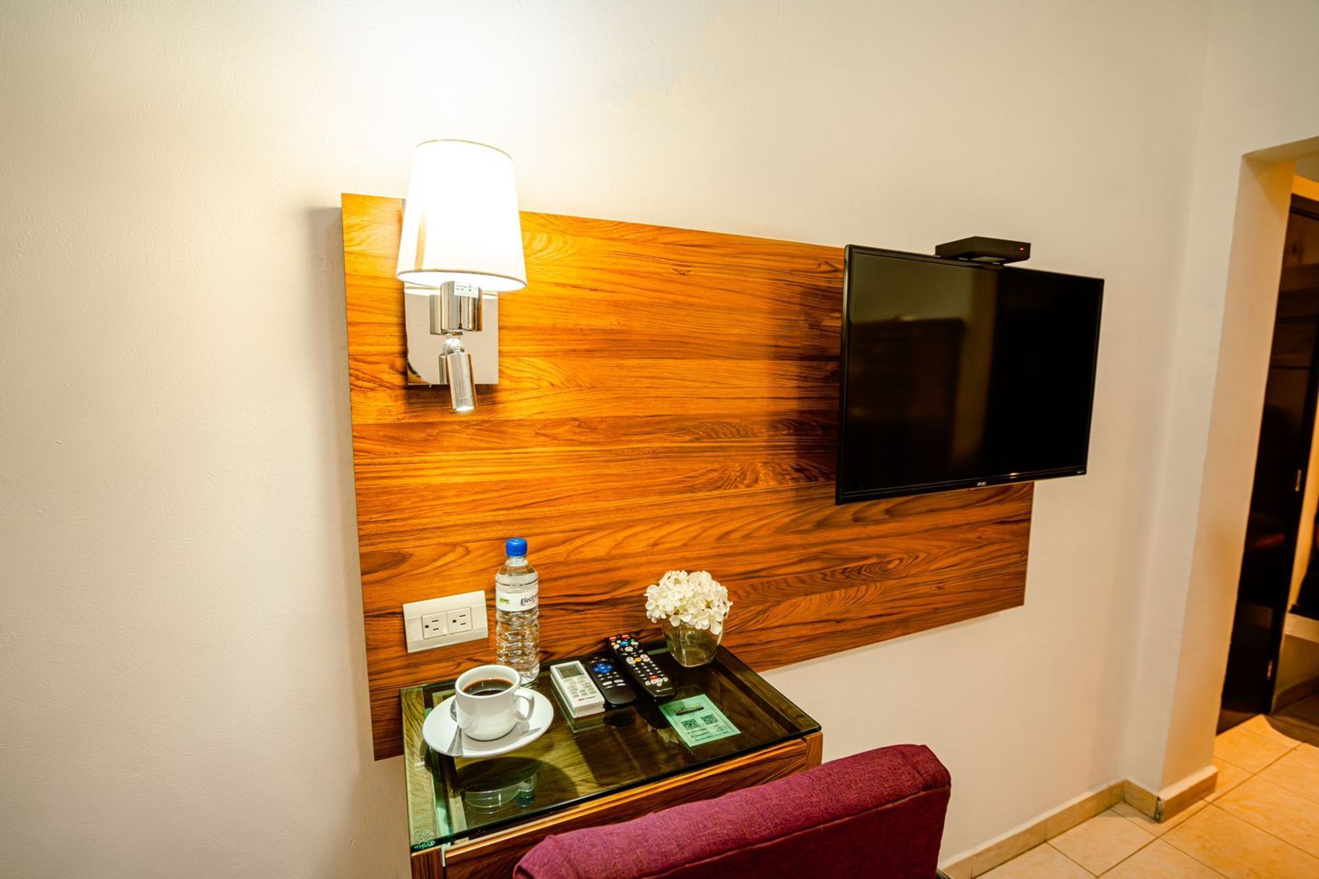 Una habitación de hotel con un televisor colgado, una lámpara y una mesa con refrescos, frente a un panel de madera.