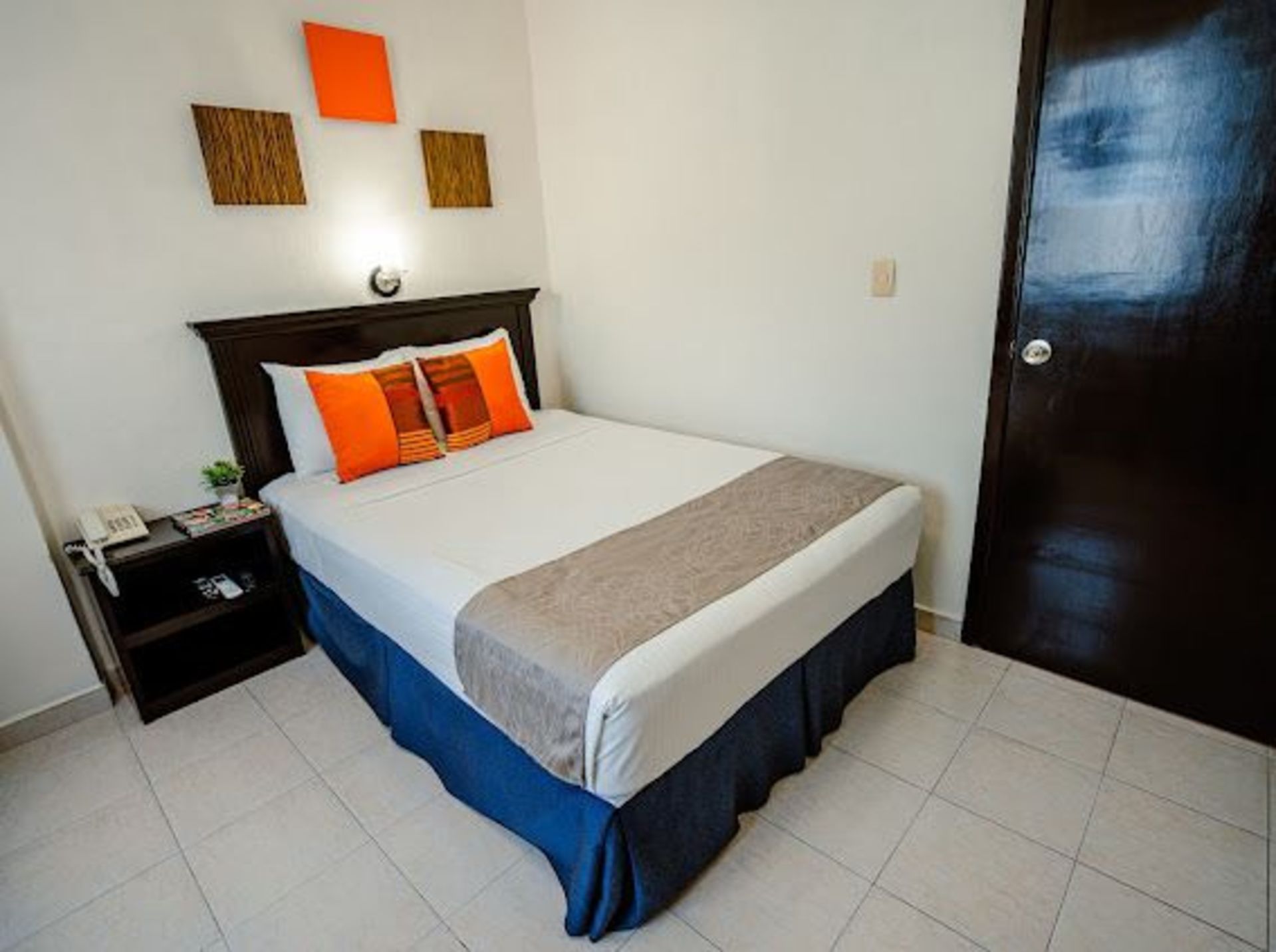 Dormitorio con cama, mesita de noche y puerta marrón oscuro. Paredes blancas, suelo de baldosas