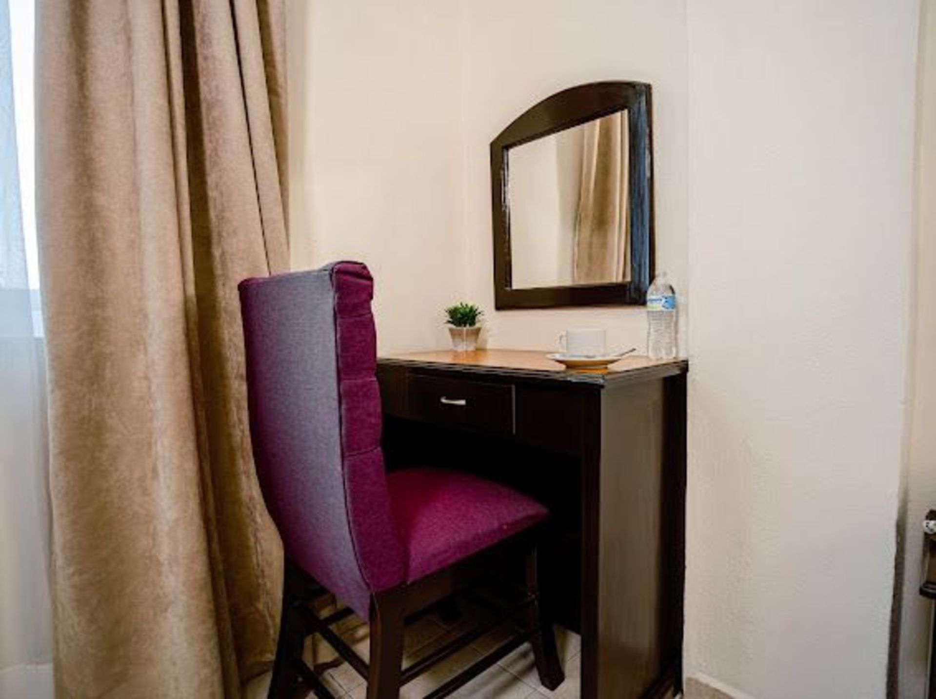 Tocador con espejo, silla color burdeos, cortinas marrones y una pequeña planta en maceta sobre el escritorio.
