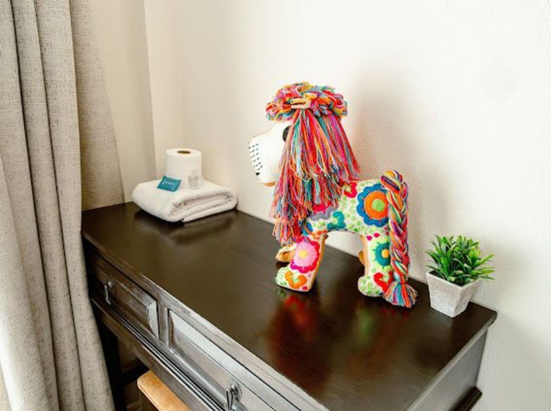 Un perro de tela decorativo sobre una cómoda de madera oscura con toallas, papel higiénico y una pequeña planta.