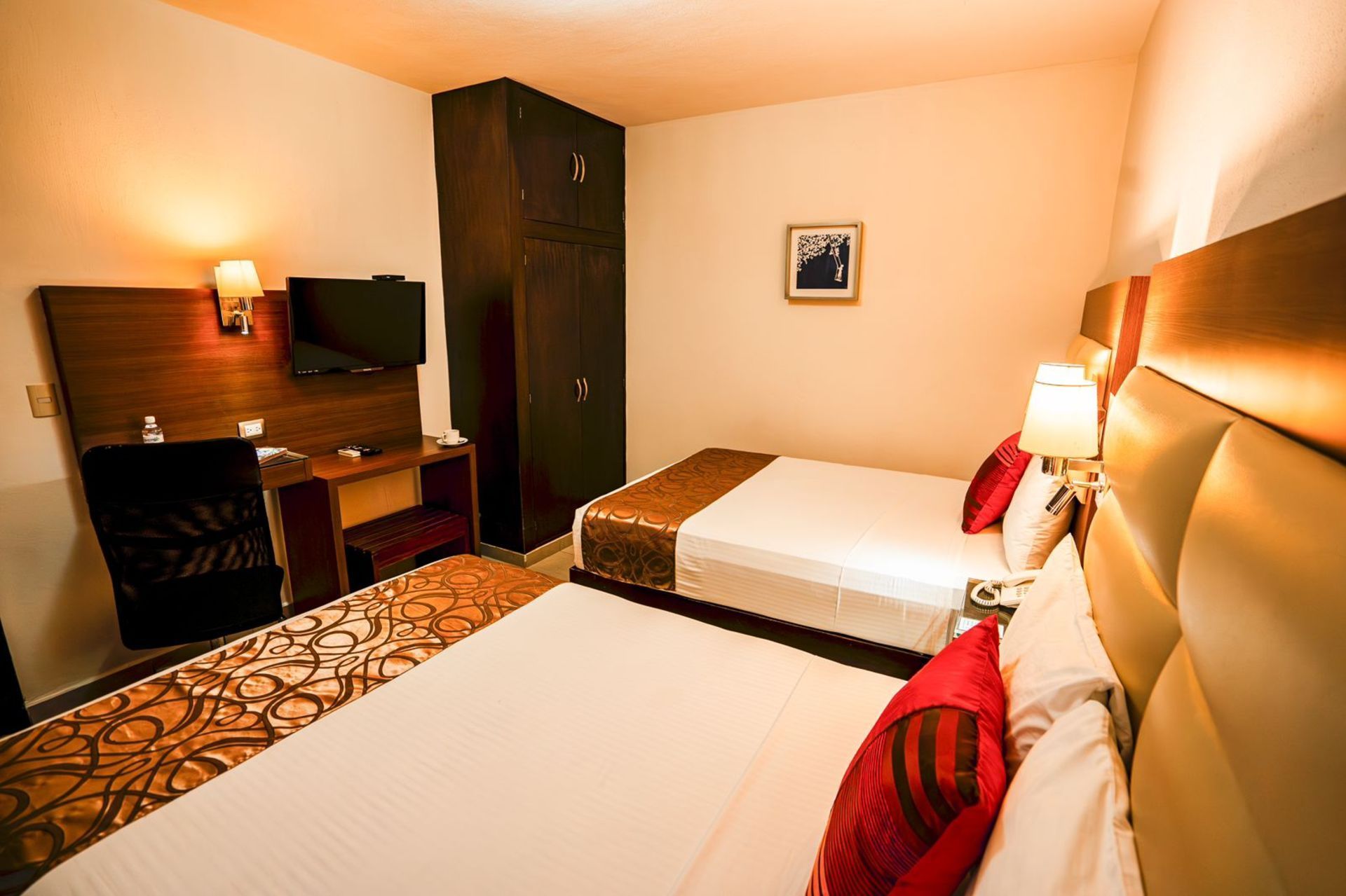 Habitación de hotel con dos camas, escritorio con TV y armario. Paredes color crema con cojines rojos.