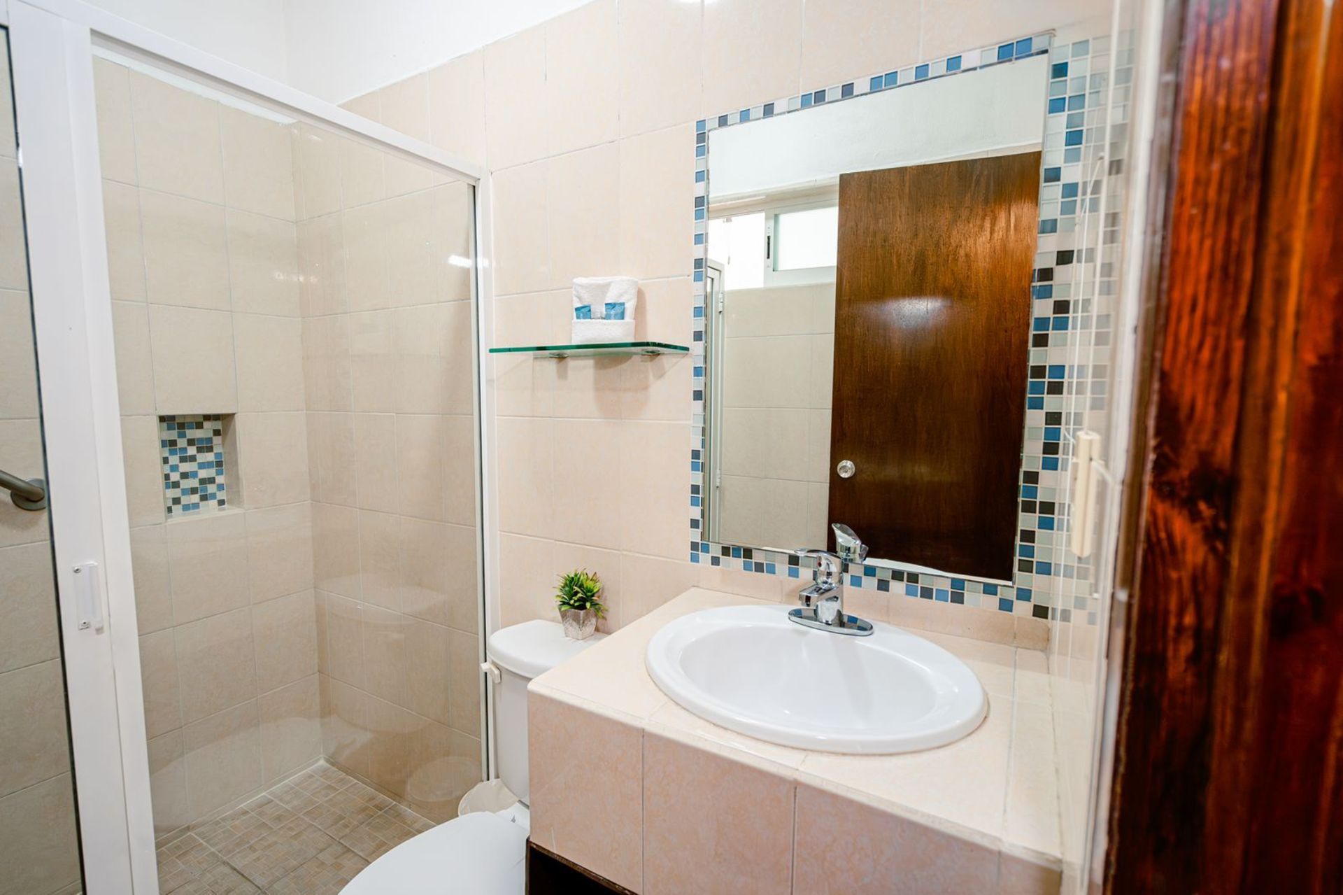 Baño con lavabo, espejo, ducha e inodoro blancos. Paredes beige con detalles de azulejos azules.