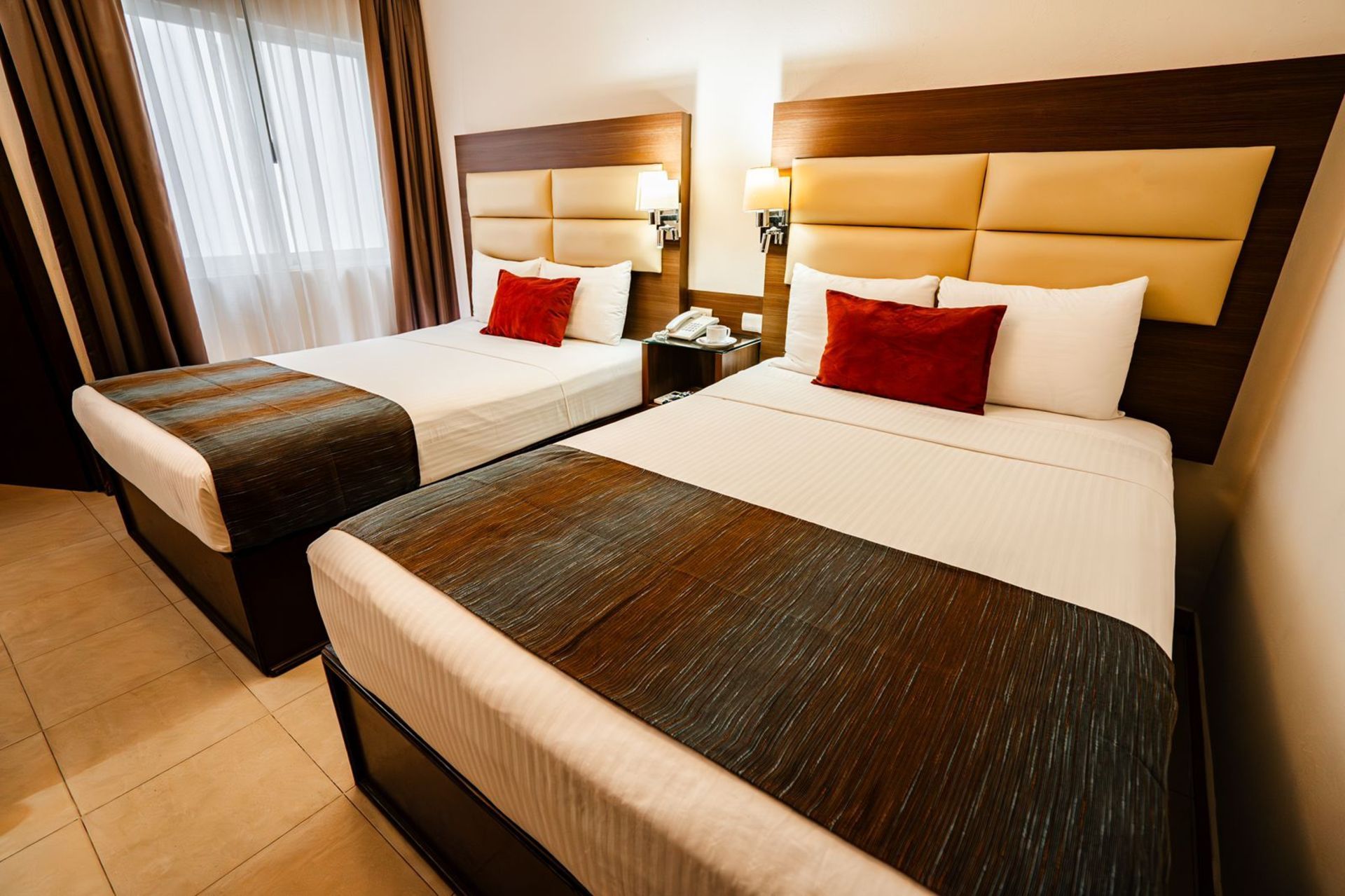 Dos camas individuales en una habitación de hotel, con cabeceros marrones y almohadas decorativas de color rojo.