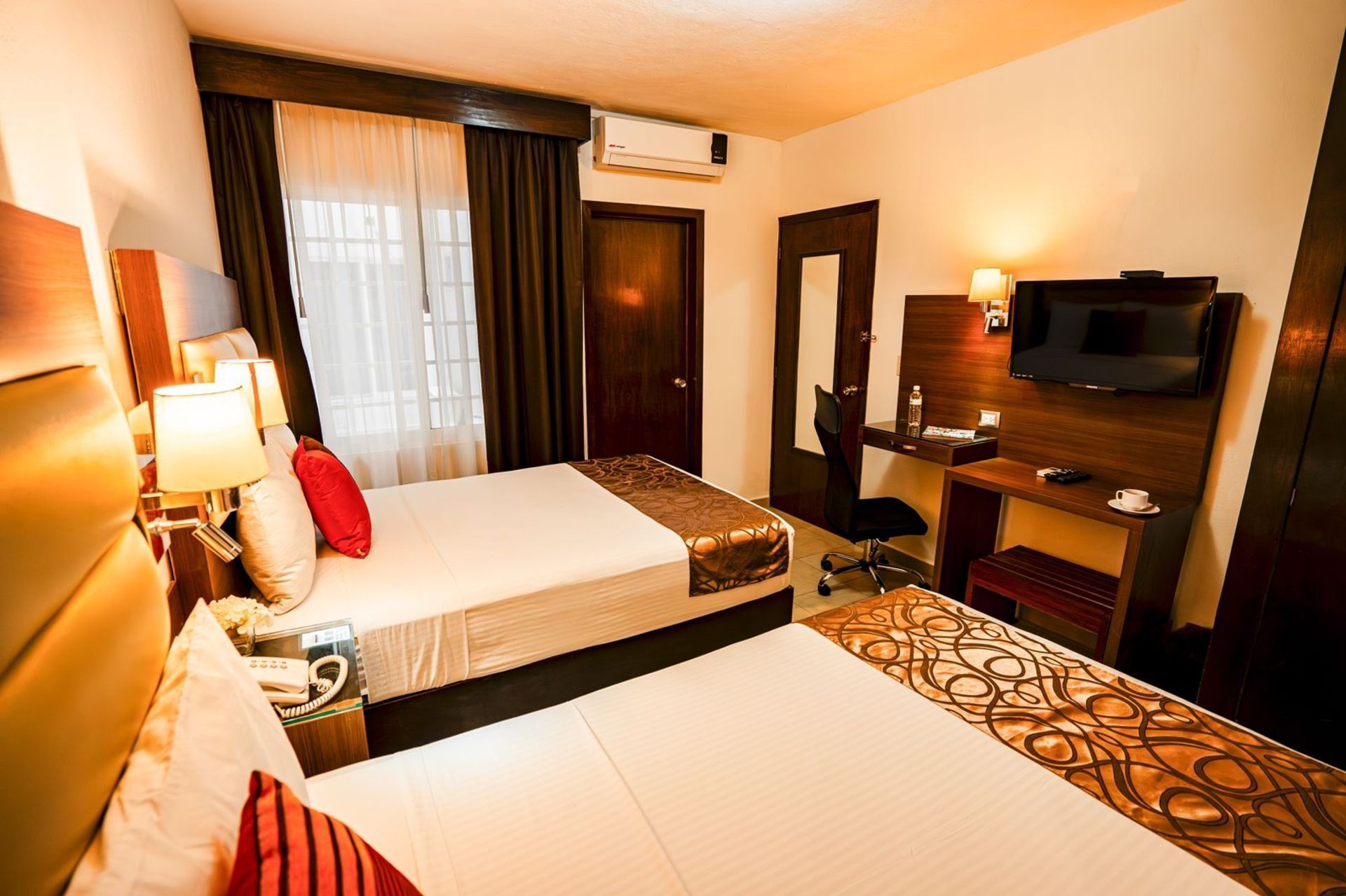 Habitación de hotel con dos camas individuales, escritorio, TV y detalles en madera oscura.