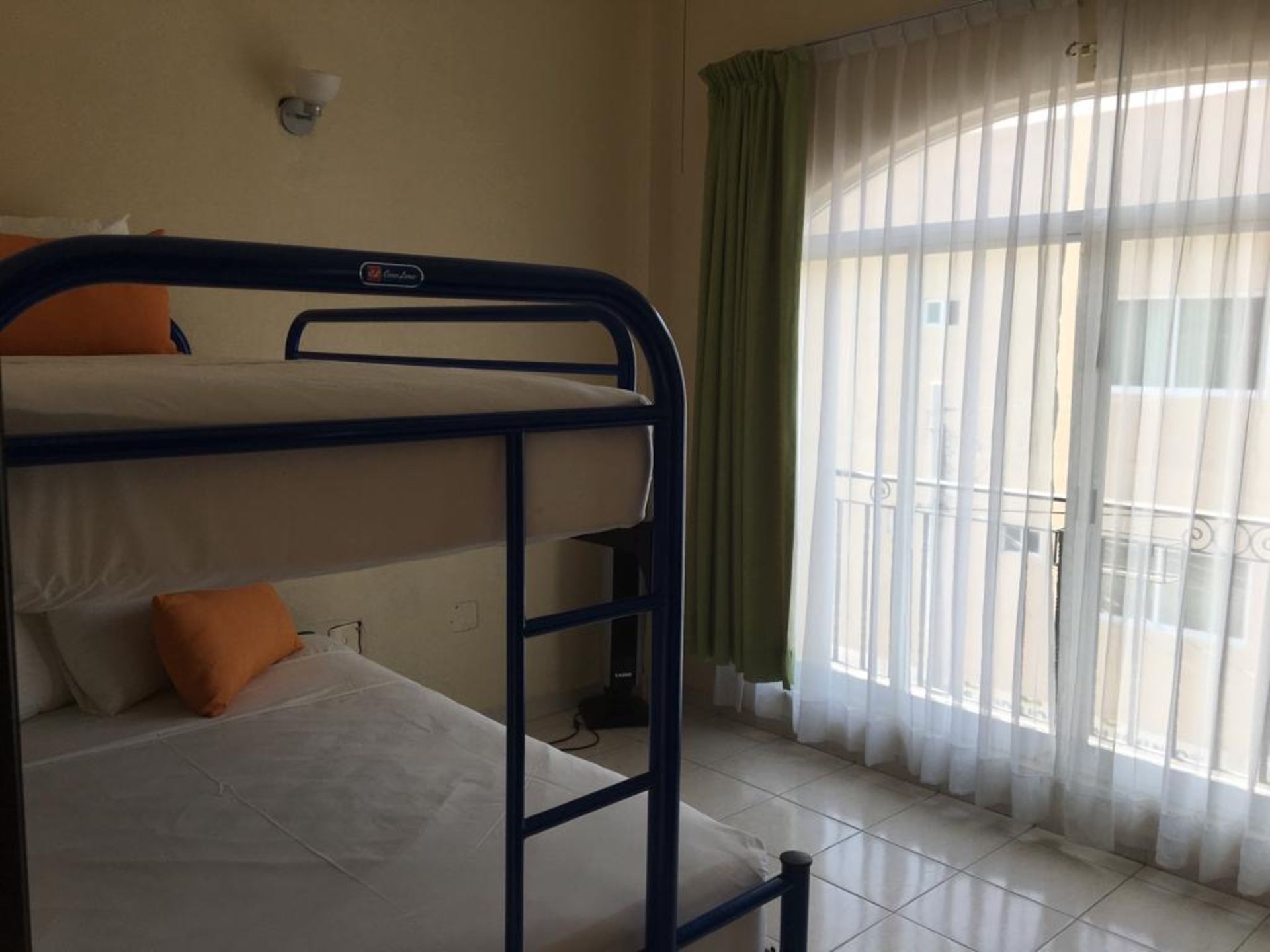 Literas azules en una habitación con ventana a un balcón. Cortina verde y ropa de cama blanca.