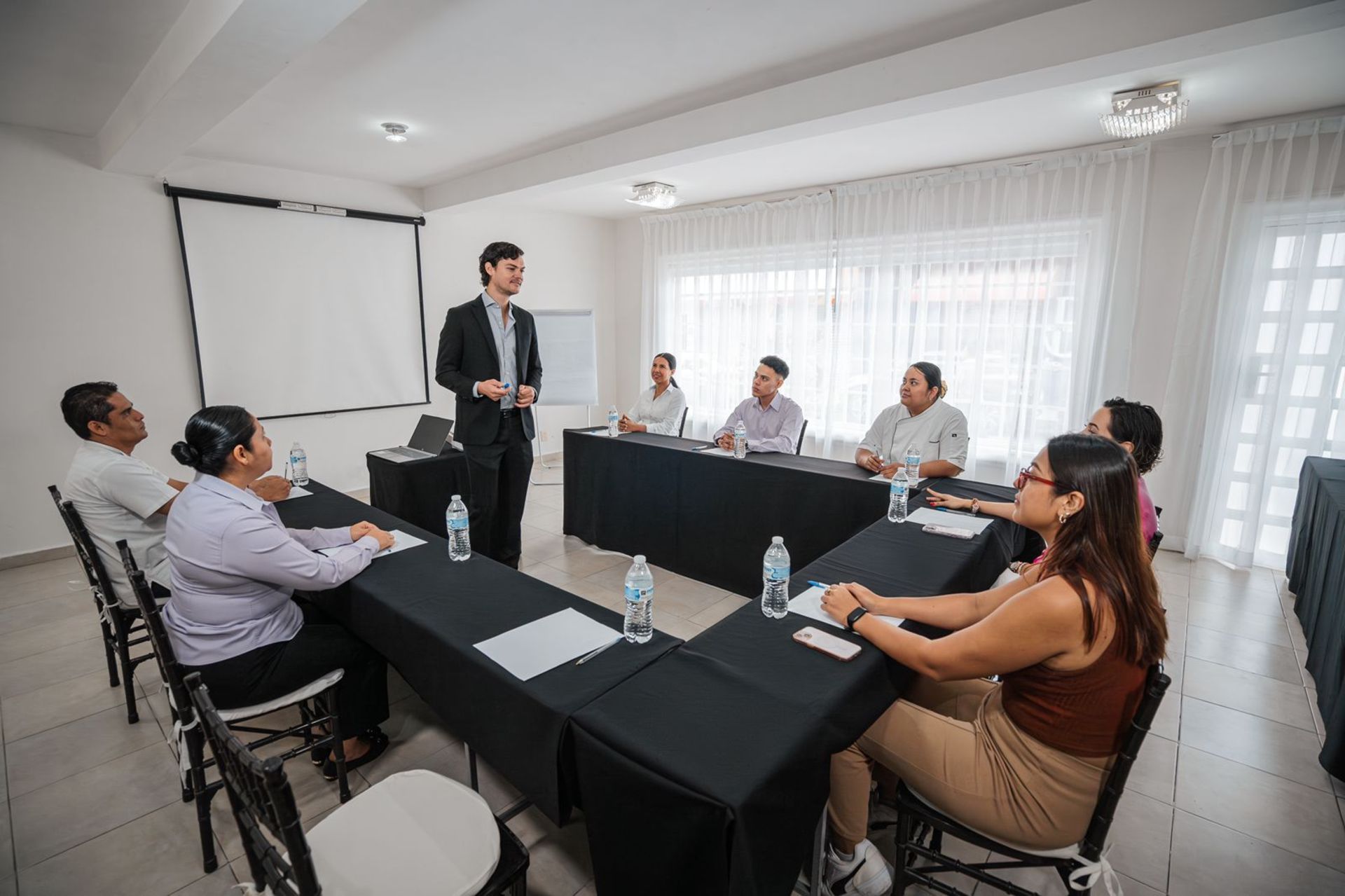 Hombre haciendo una presentación ante un pequeño grupo sentado alrededor de una mesa en forma de U en una habitación luminosa