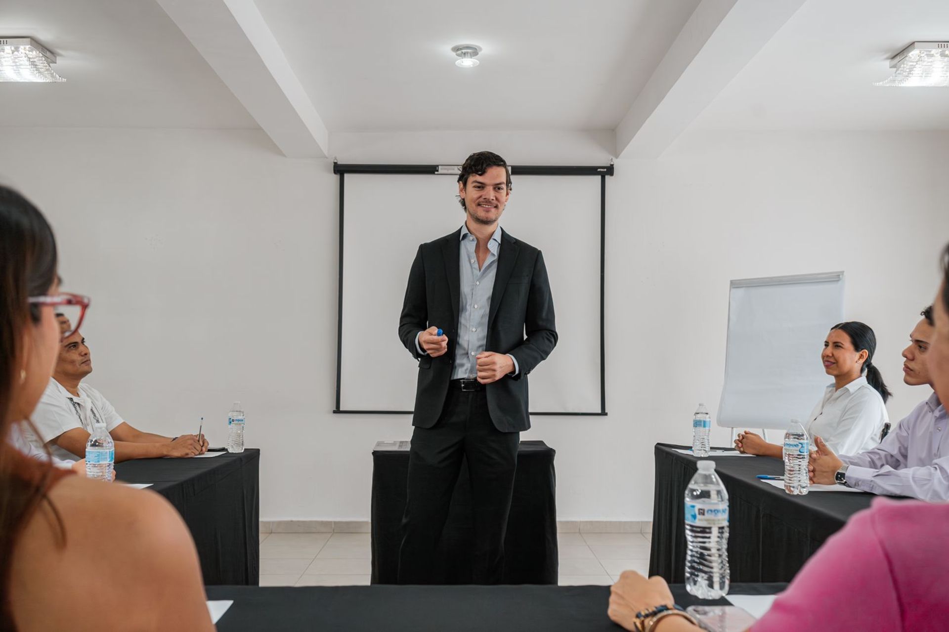 Hombre de traje dando una presentación en una sala de conferencias. Asistentes sentados en mesas.