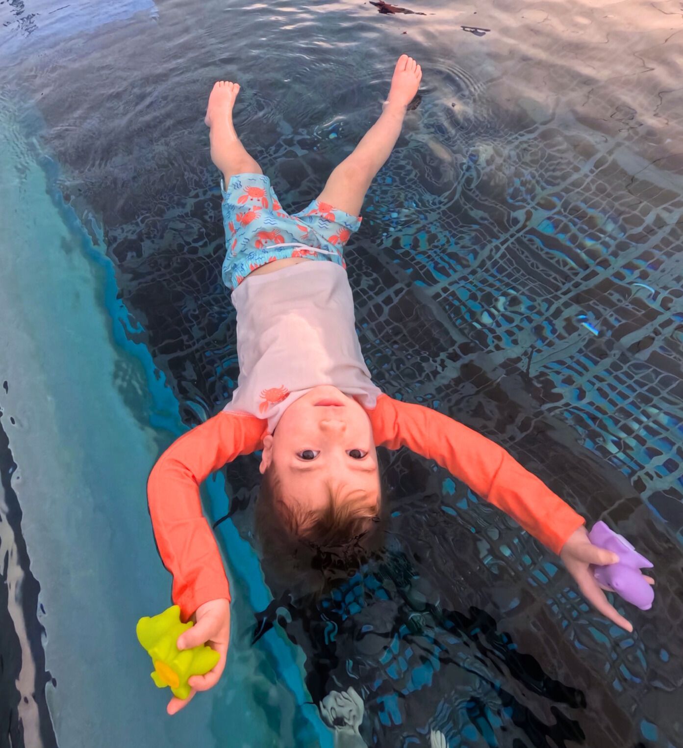 Child floating in pool, wearing rash guard and swim trunks, holding toys, looking up.