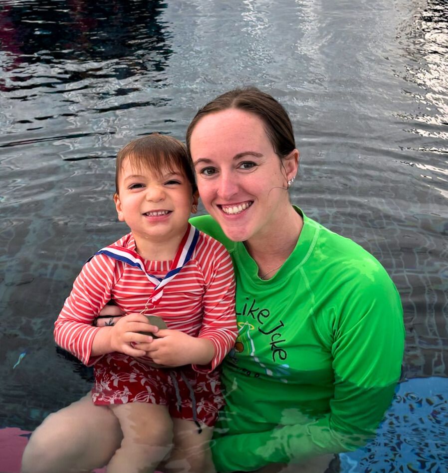 Woman in green shirt and young child in a swimsuit smiling in a swimming pool.