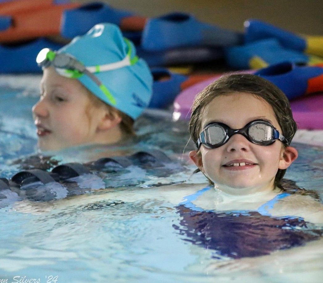 Two girls in a swimming pool; one smiling with goggles, the other with a swim cap and goggles.