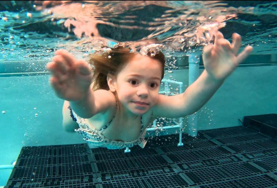 Young girl swimming underwater, arms outstretched, smiling in a blue pool.