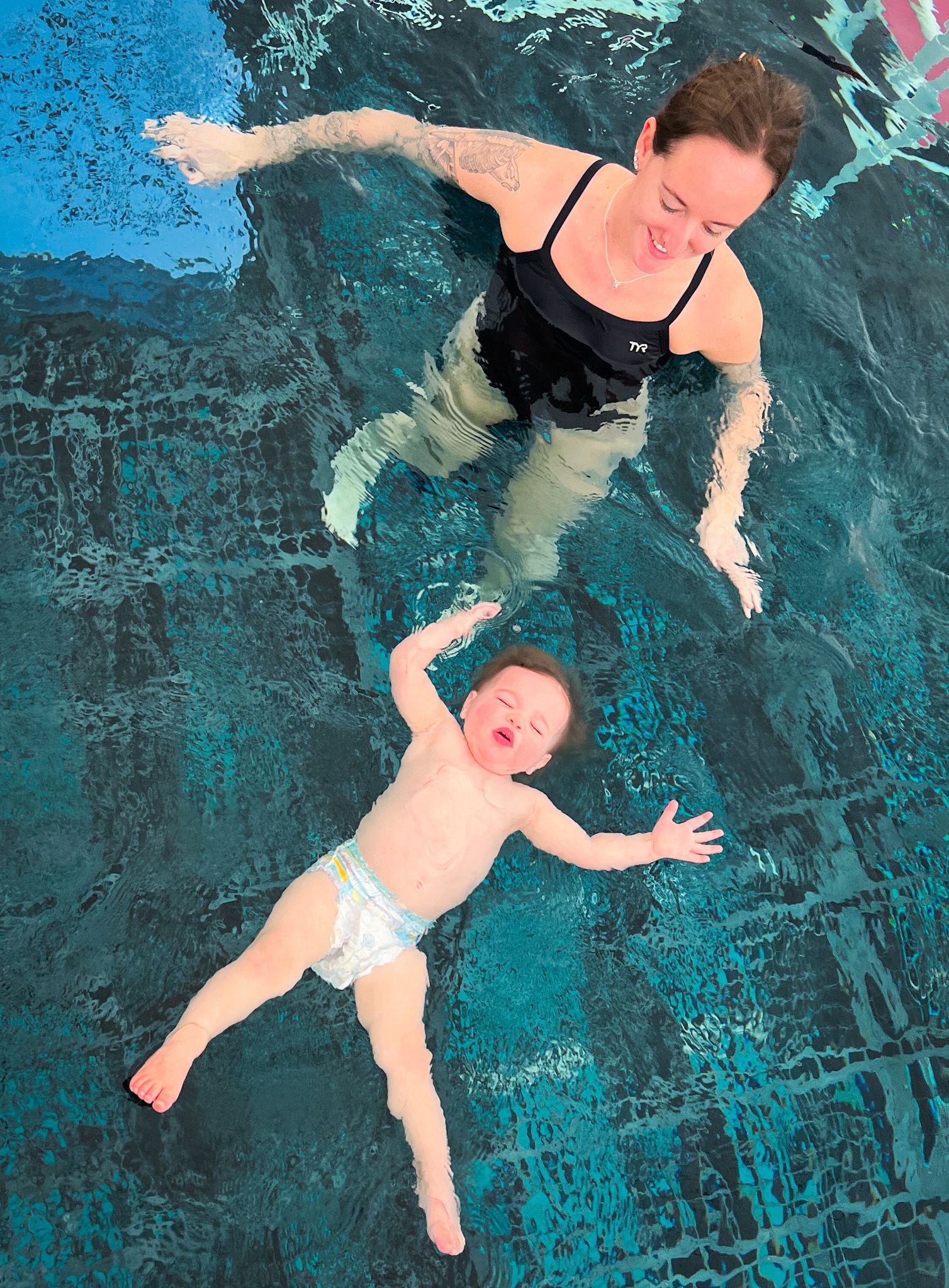 Woman in black swimsuit and baby in diaper floating in a pool, baby with mouth open.