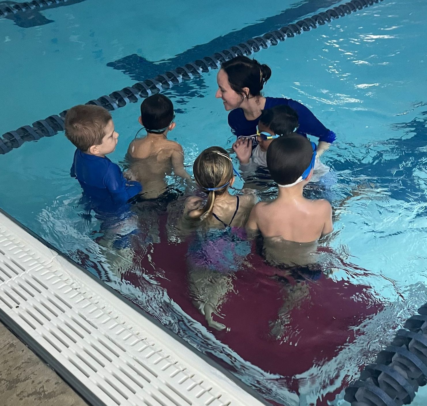 Swimming instructor with six children in a pool, smiling. Children are in swimwear.