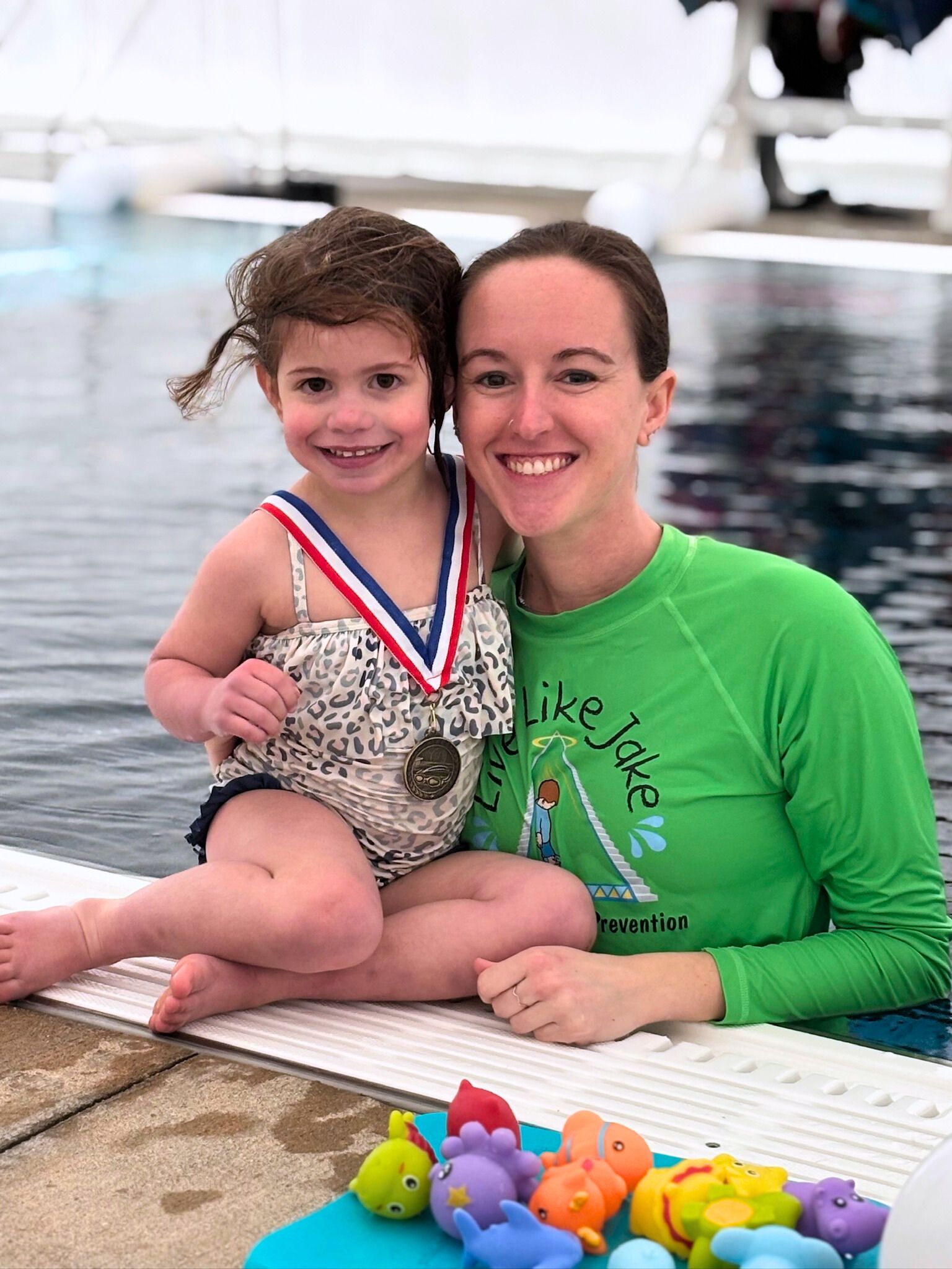 Woman in green shirt with young girl in swimsuit by a pool, both smiling. Girl wears medal.