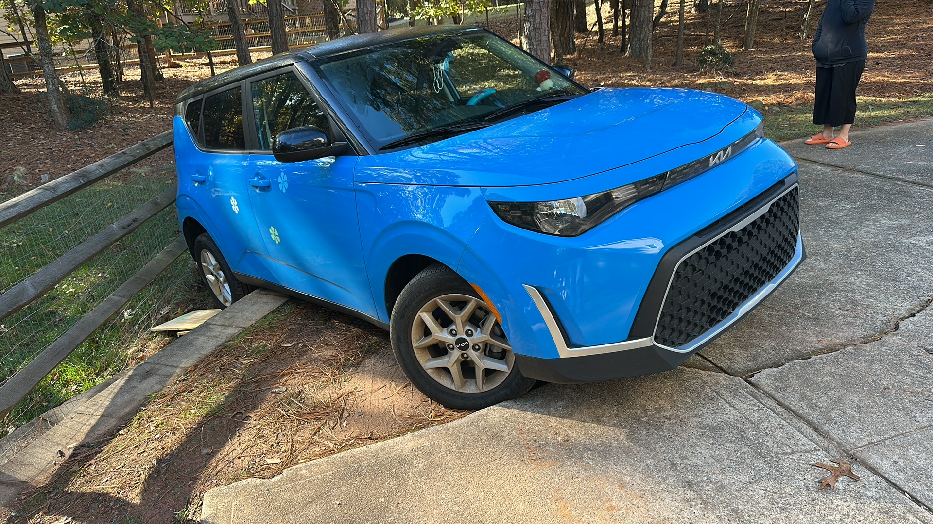 A bright blue Kia Soul is angled off a concrete driveway, resting against a wooden post-and-rail fence.