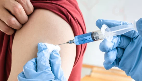 Person receiving a blue liquid injection in their upper arm; a gloved hand holds the syringe, while another hand holds a cotton swab.
