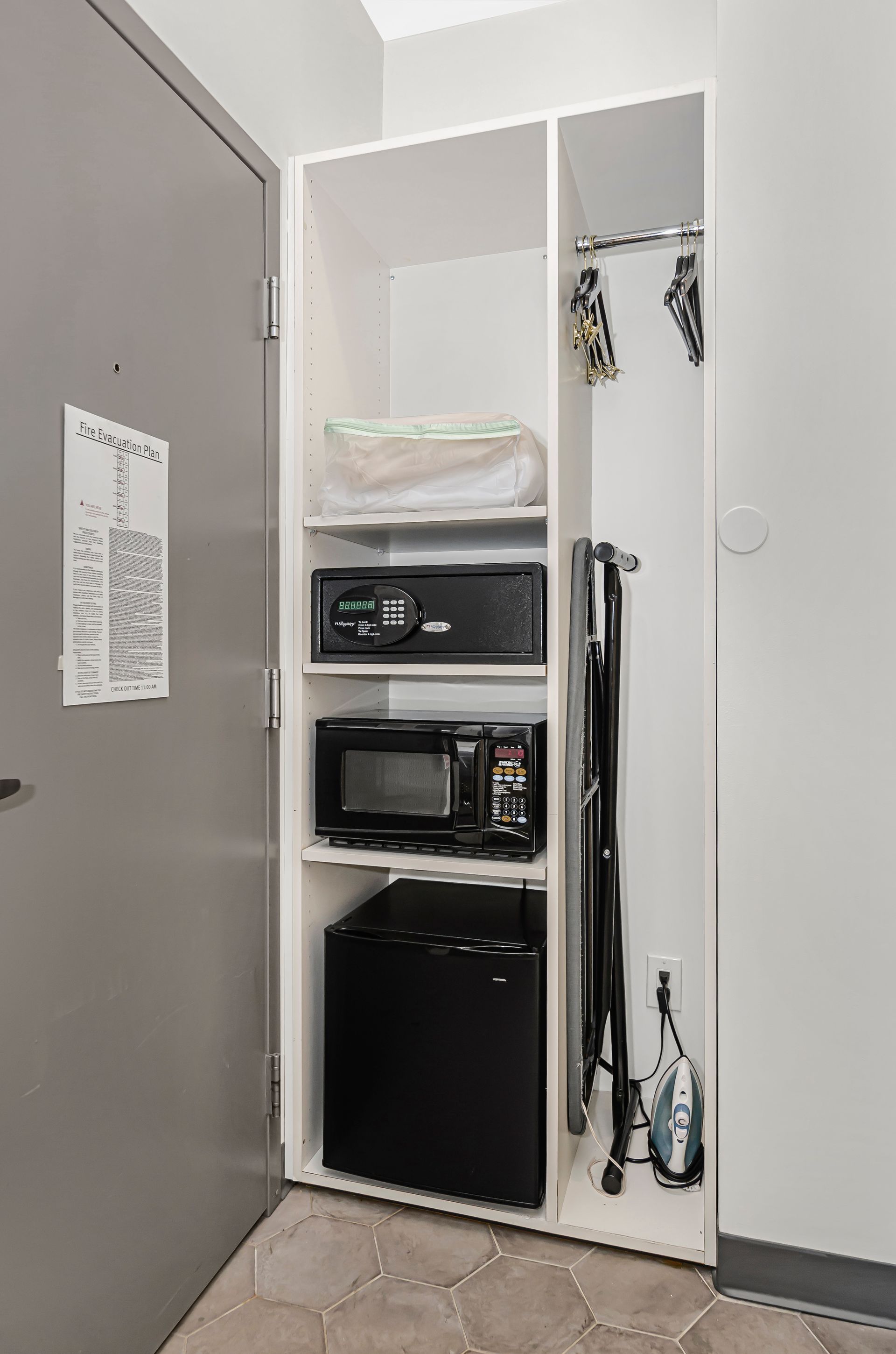 A tall, white open cabinet in a room holding a white package, black safe, microwave, mini-fridge, iron, and ironing board.